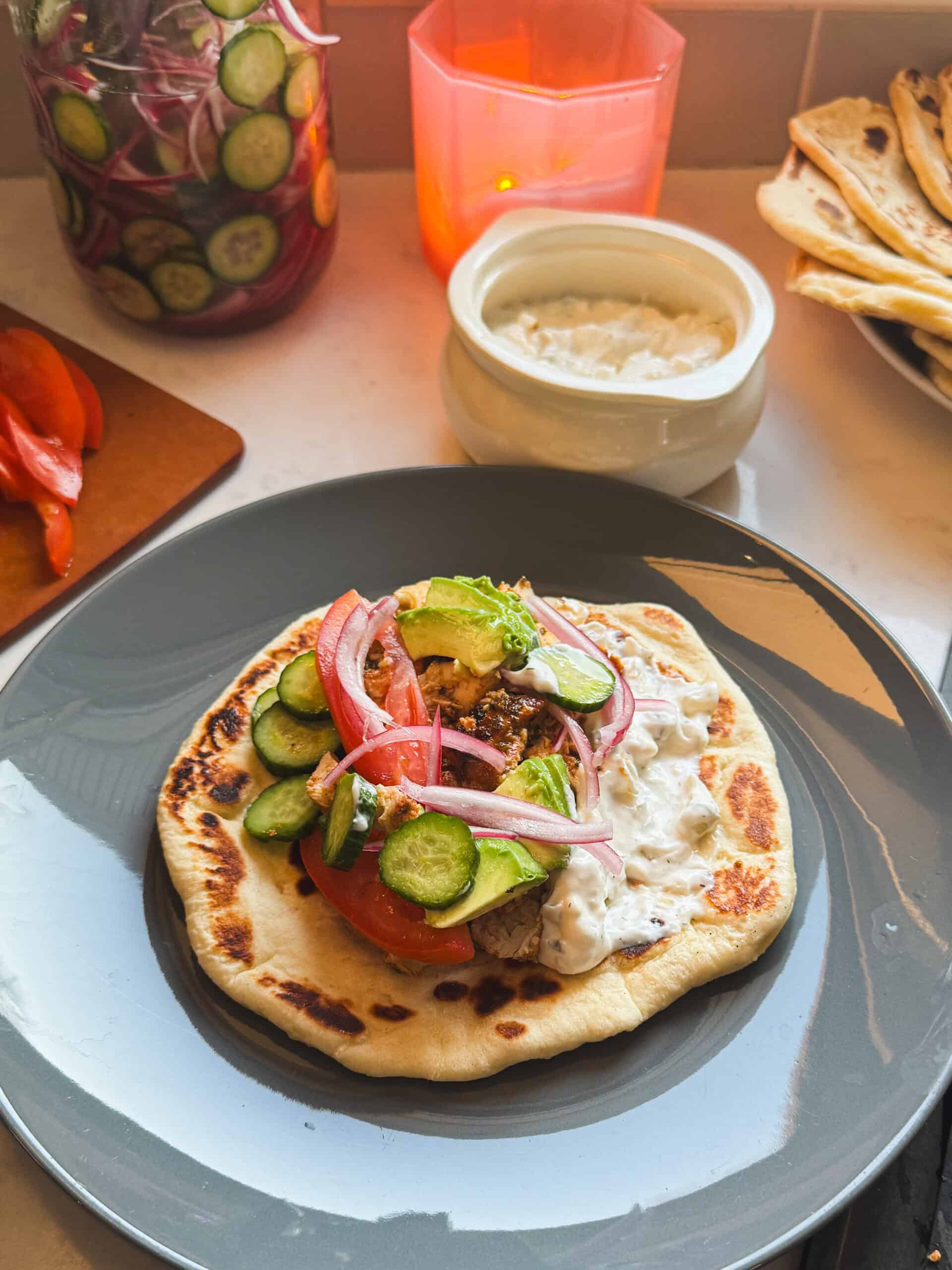 Chicken gyro sandwich being assembled on soft sweet flatbread with cucumbers, red onions, tomatoes, and avocado