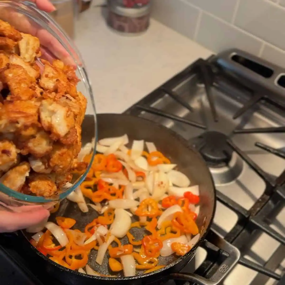 Fried chicken cubes being poured into a pan of sautéed onions and peppers.