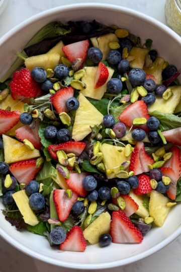 Fresh strawberry poppyseed salad with power greens, strawberries, blueberries, pineapple, and pistachios in a large bowl.