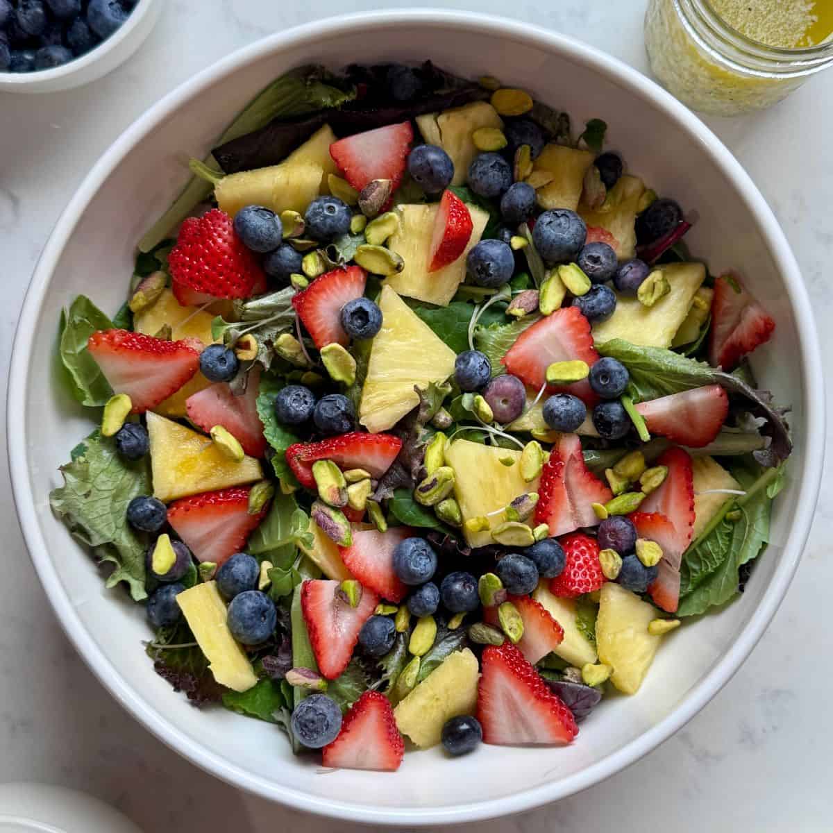 Fresh strawberry poppyseed salad with power greens, strawberries, blueberries, pineapple, and pistachios in a large bowl.