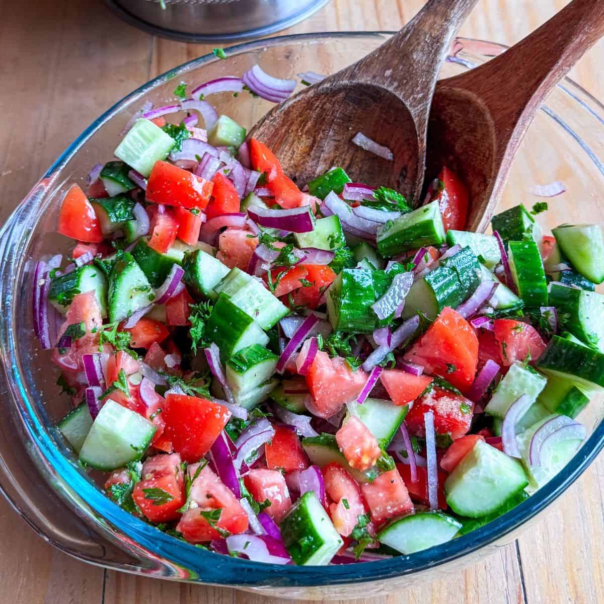 Glass bowl of Mediterranean cucumber tomato salad with fresh herbs and wooden serving tongs.