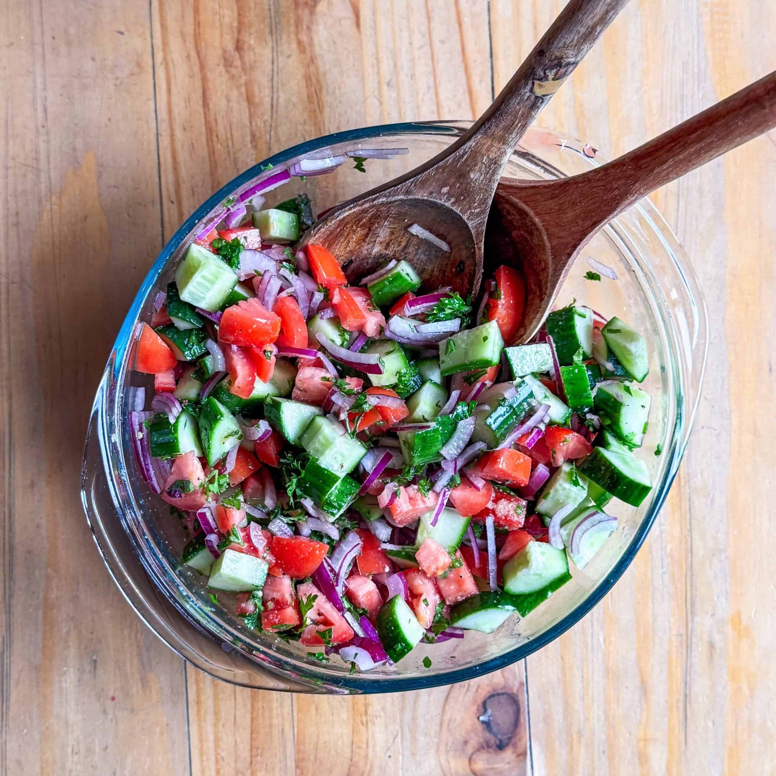 Glass bowl of Mediterranean cucumber tomato salad with fresh herbs and wooden serving tongs.