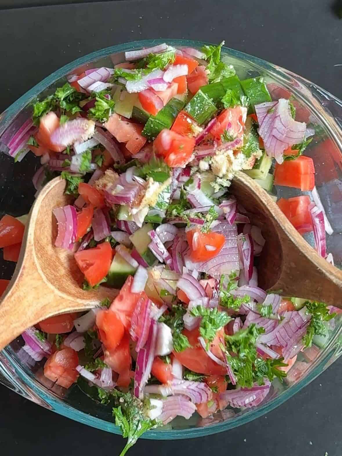 Mediterranean cucumber tomato salad being mixed with dressing and spices in a glass bowl.