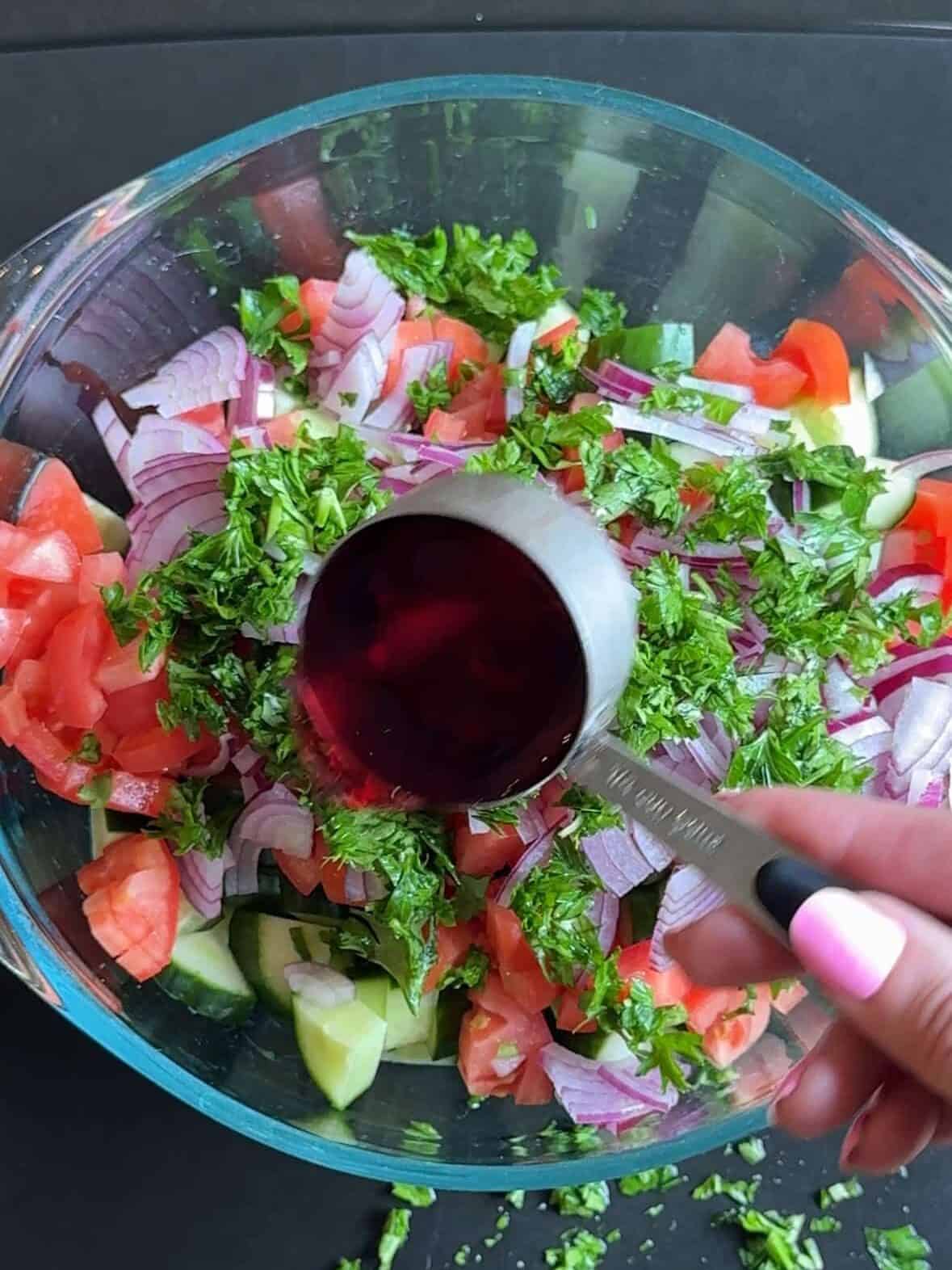 Red wine vinegar, olive oil, and spices being added to cucumber and tomato salad ingredients.