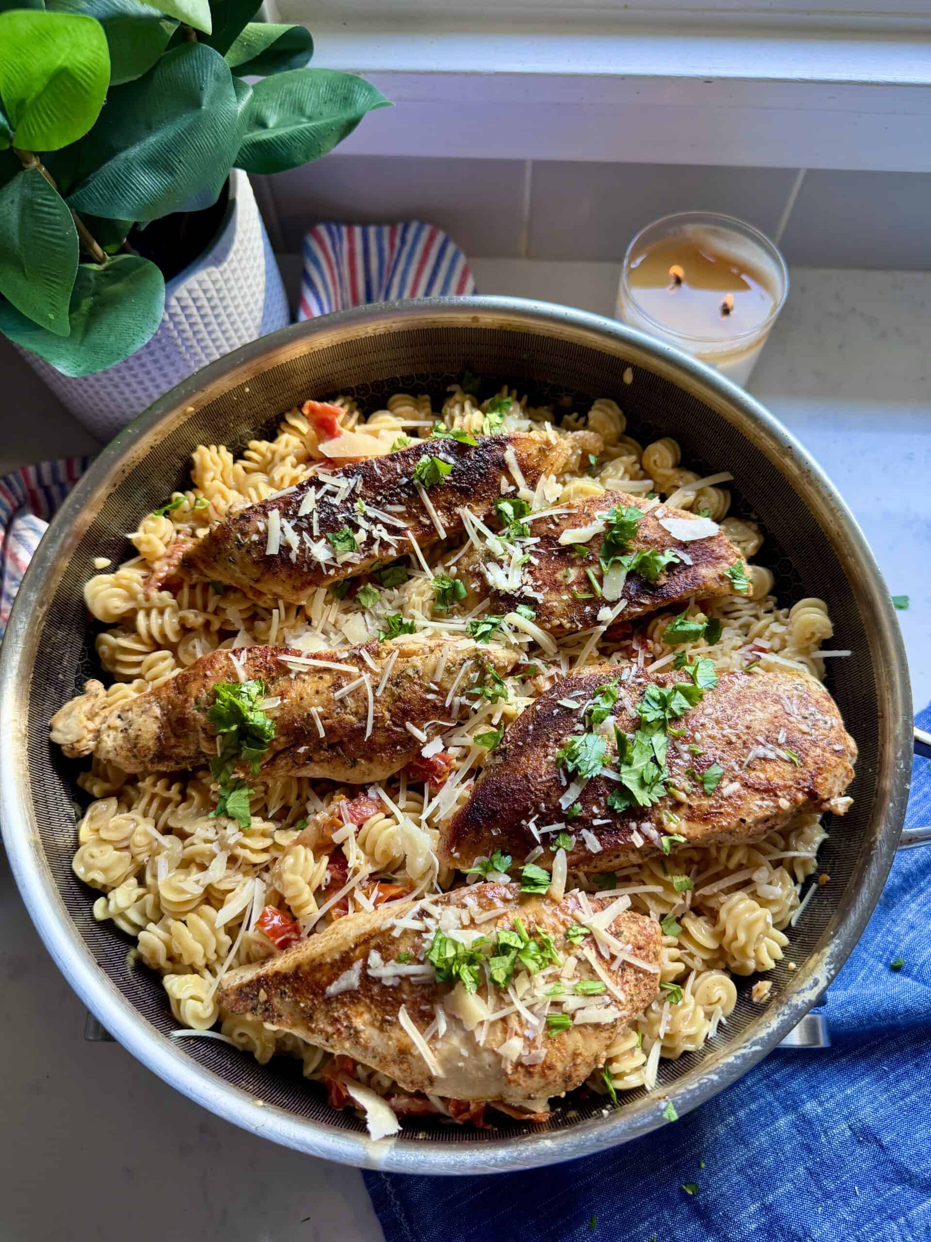 Pot of creamy sun-dried tomato chicken and pasta with candle and plant decor in the background.