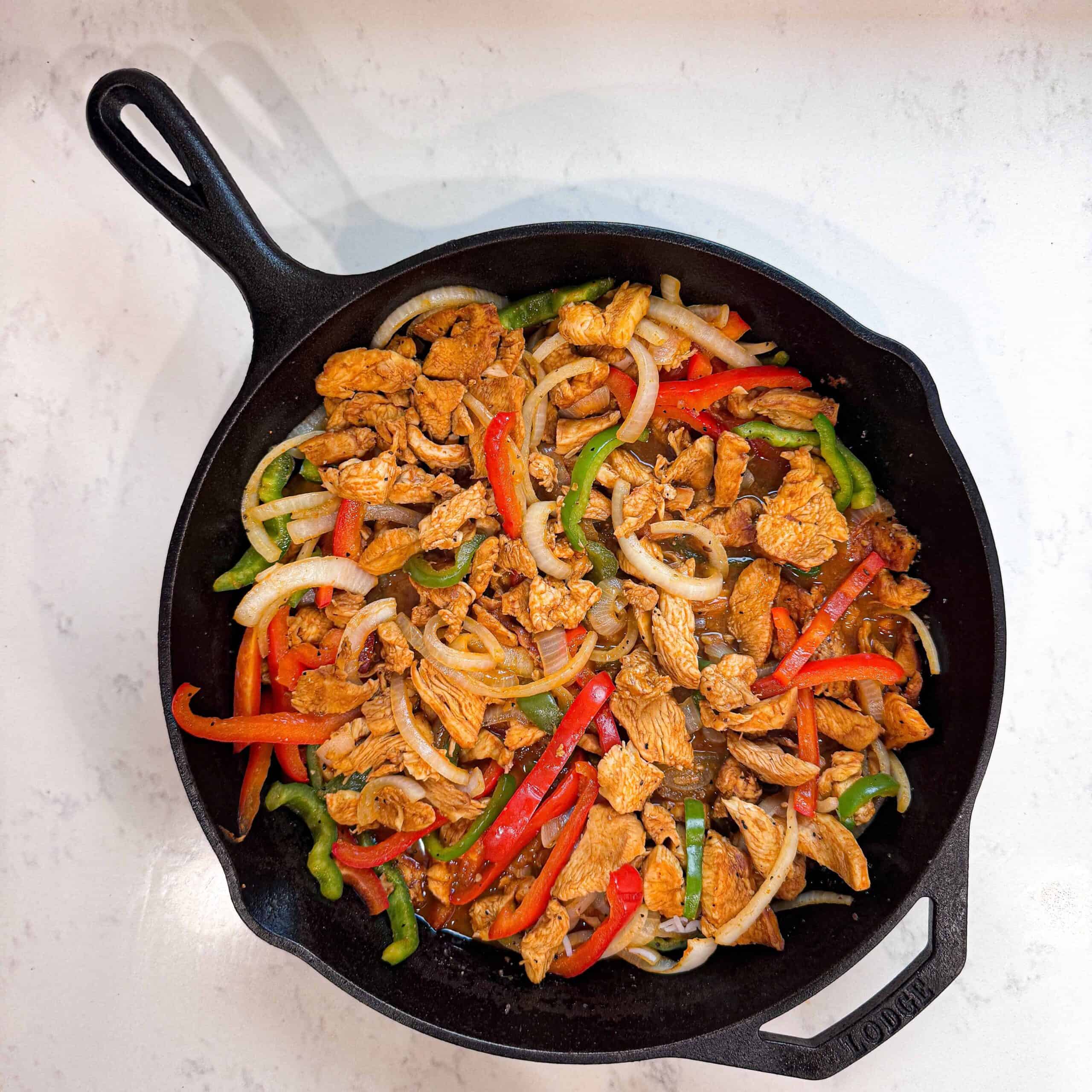 Overhead shot of chicken fajitas with onions and peppers in a cast iron skillet on a wooden surface.