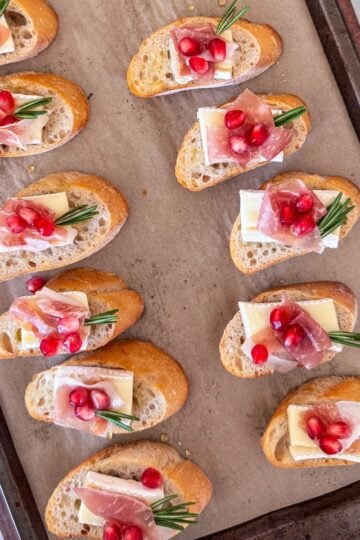 Overhead view of finished crostini with rosemary sprigs and scattered pomegranate seeds.