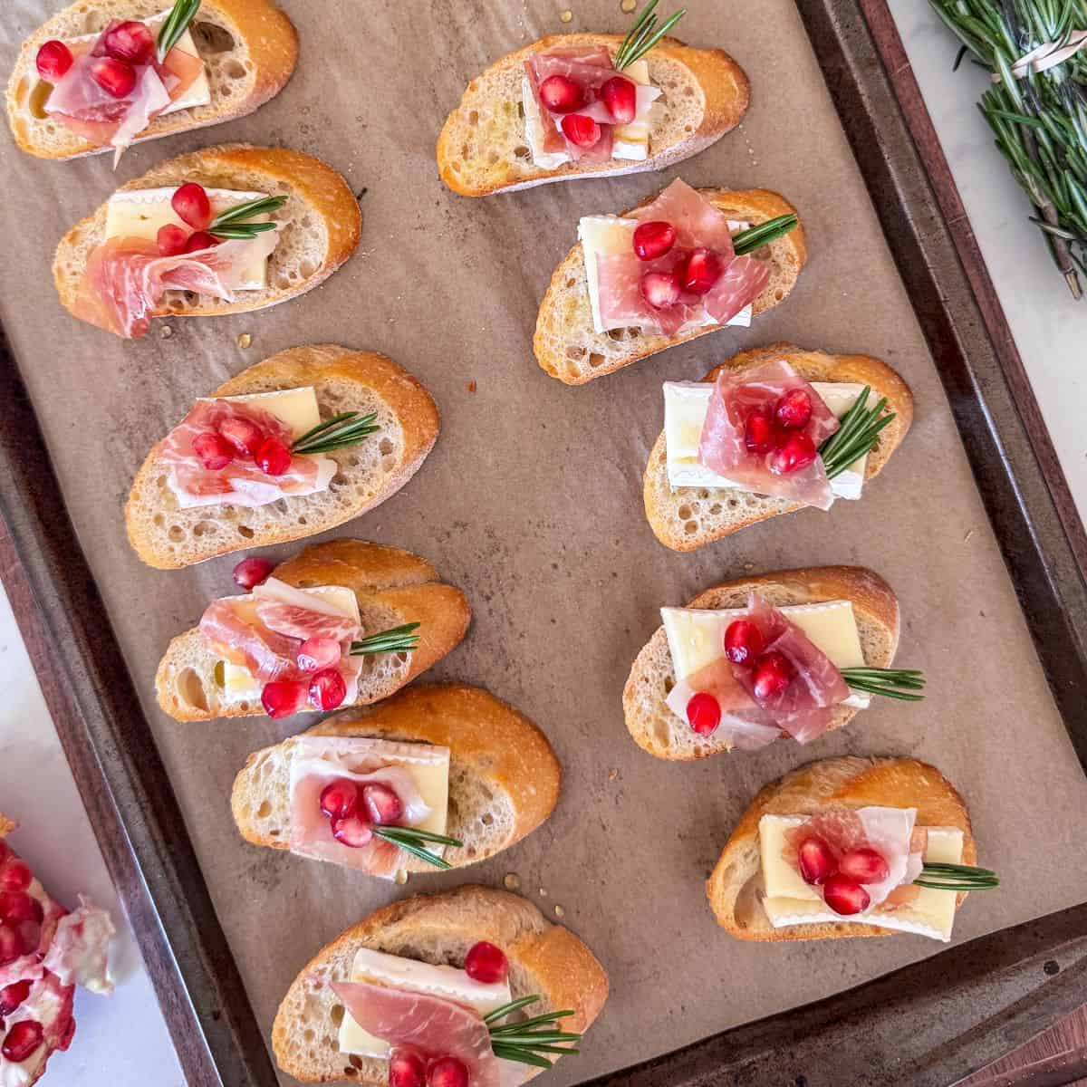 Overhead view of finished crostini with rosemary sprigs and scattered pomegranate seeds.