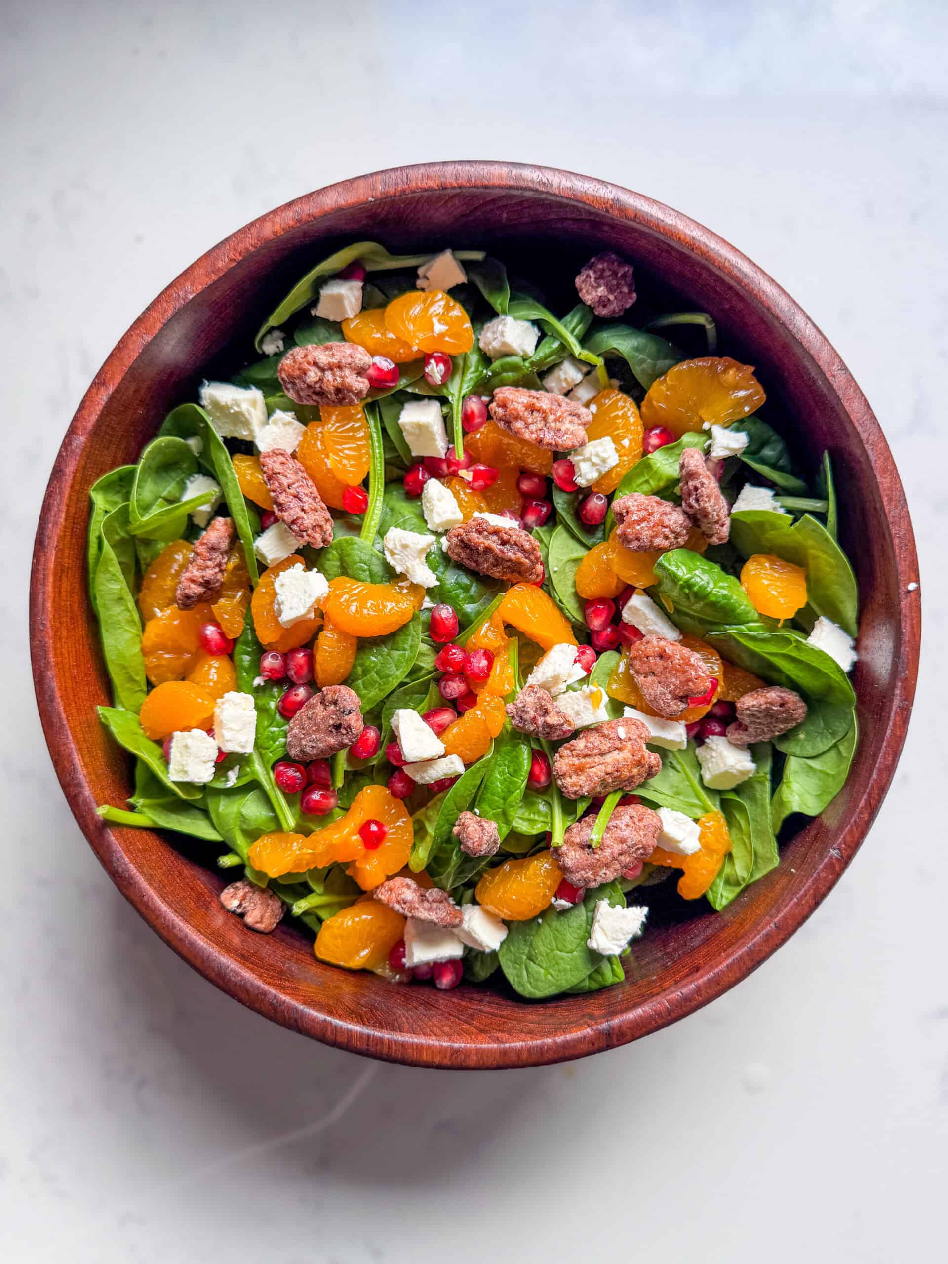 Overhead view of a feta and pomegranate salad in a wooden bowl with spinach, oranges, candied pecans, and pomegranate arils.