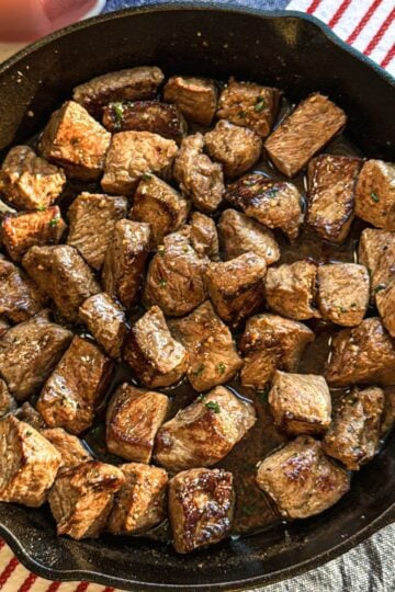 Overhead photo of garlic butter steak bites in cast iron pan on tablecloth with candle.