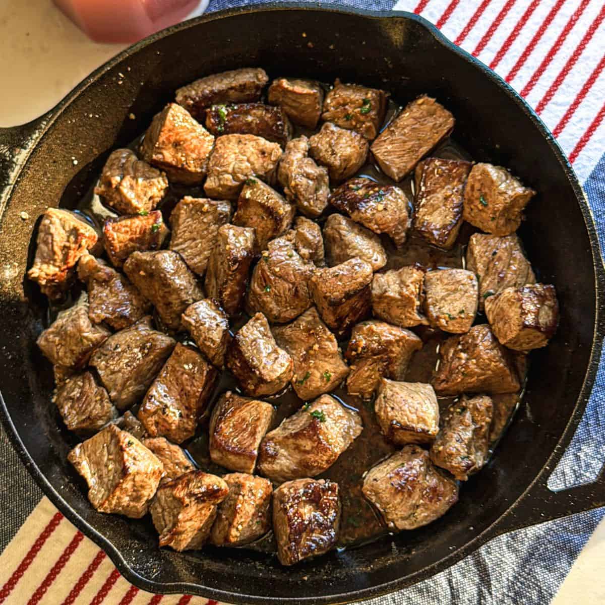 Overhead photo of garlic butter steak bites in cast iron pan on tablecloth with candle.