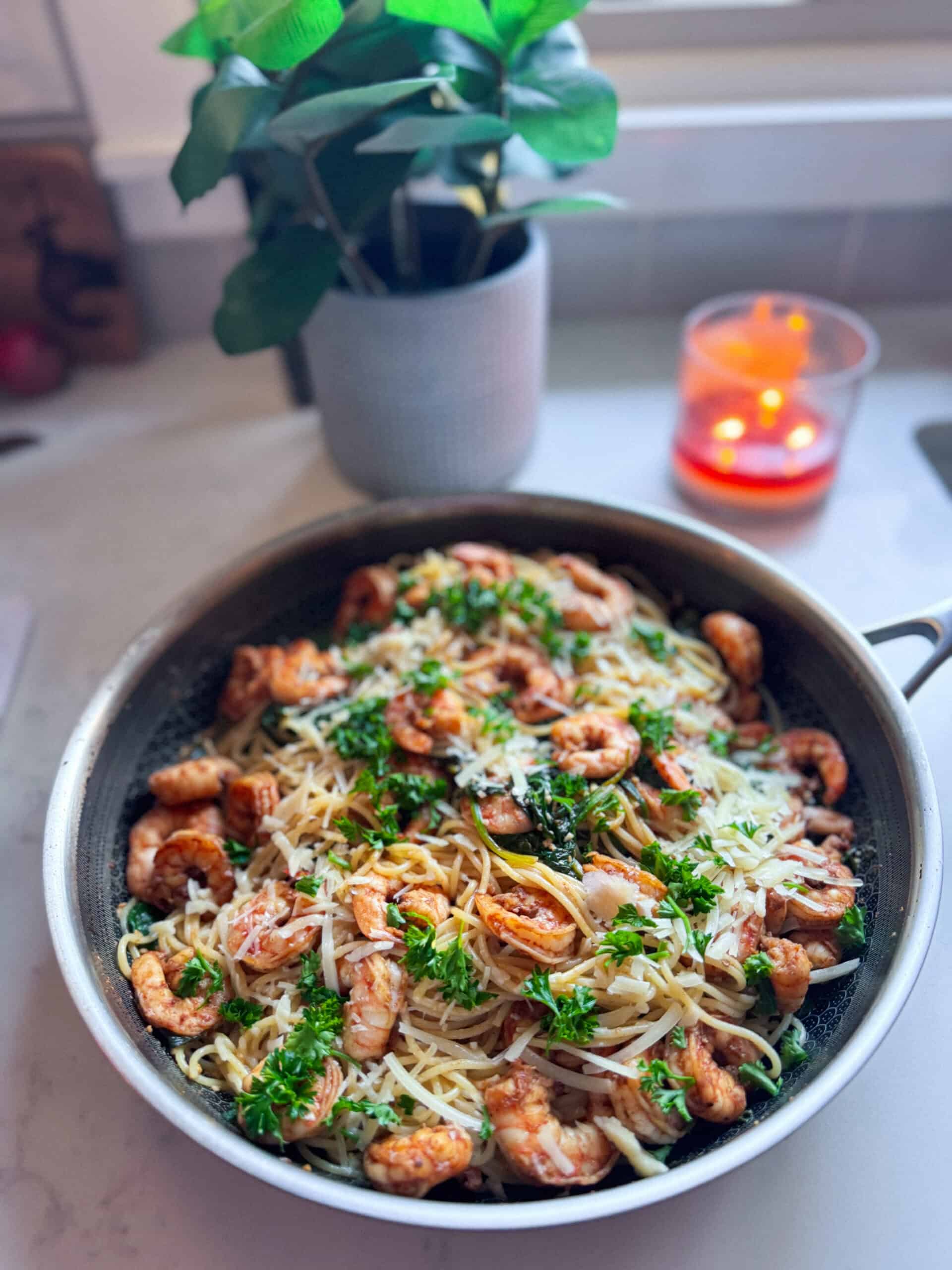 Angle view of shrimp with spinach and pasta in a skillet, with a candle and plant blurred in the background.