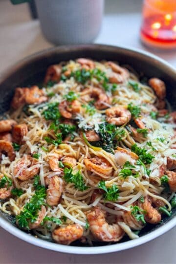 Angle view of shrimp with spinach and pasta in a skillet, with a candle and plant blurred in the background.