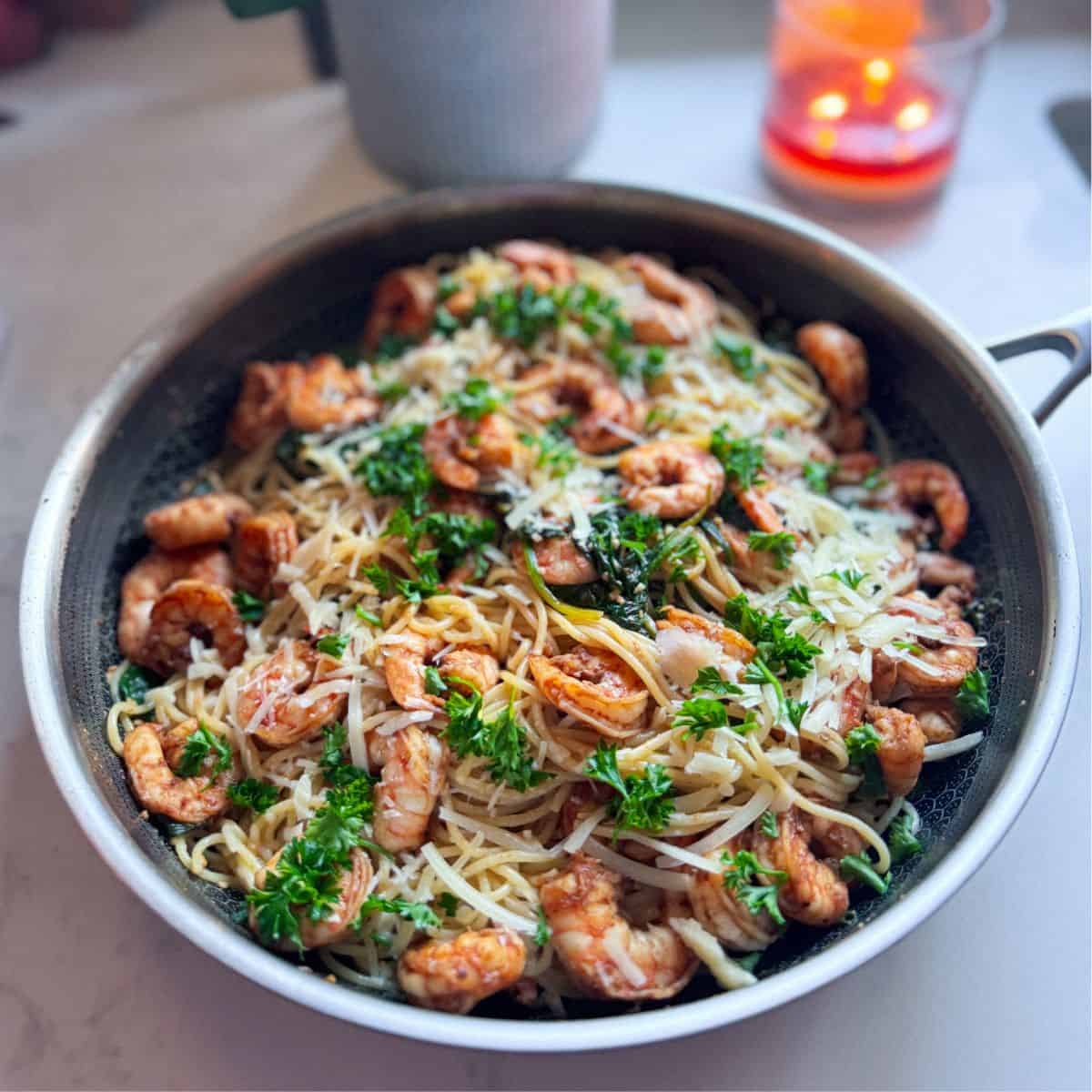 Angle view of shrimp with spinach and pasta in a skillet, with a candle and plant blurred in the background.