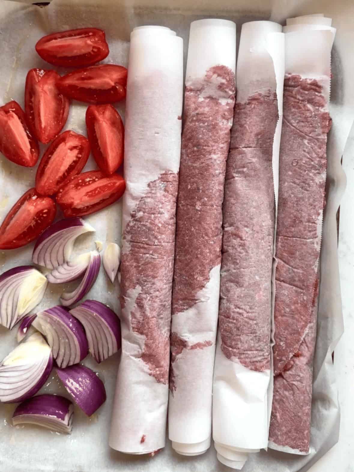 Rolled gyro meat in parchment on a sheet pan with onions and tomatoes