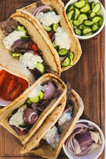 Gyro sandwiches with ground beef gyro meat, tzatziki, tomatoes, onions, and cucumbers on a wooden board