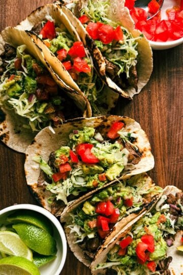 Overhead view of steak street tacos made with marinated steak, slaw, guacamole, and diced tomatoes on a wooden cutting board