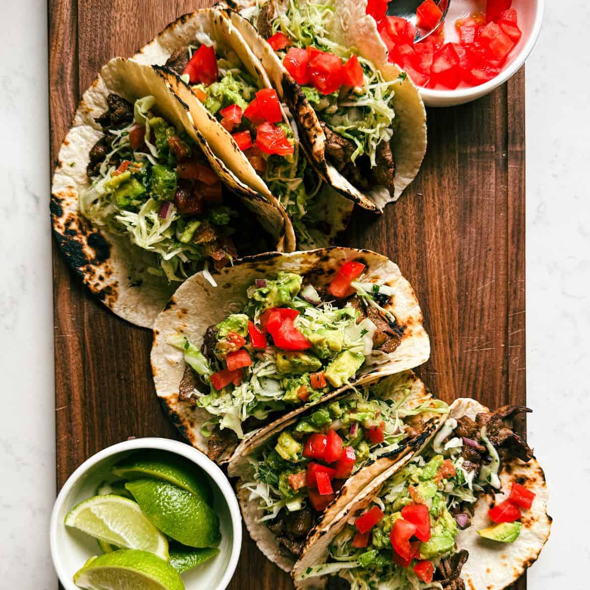 Overhead view of steak street tacos made with marinated steak, slaw, guacamole, and diced tomatoes on a wooden cutting board