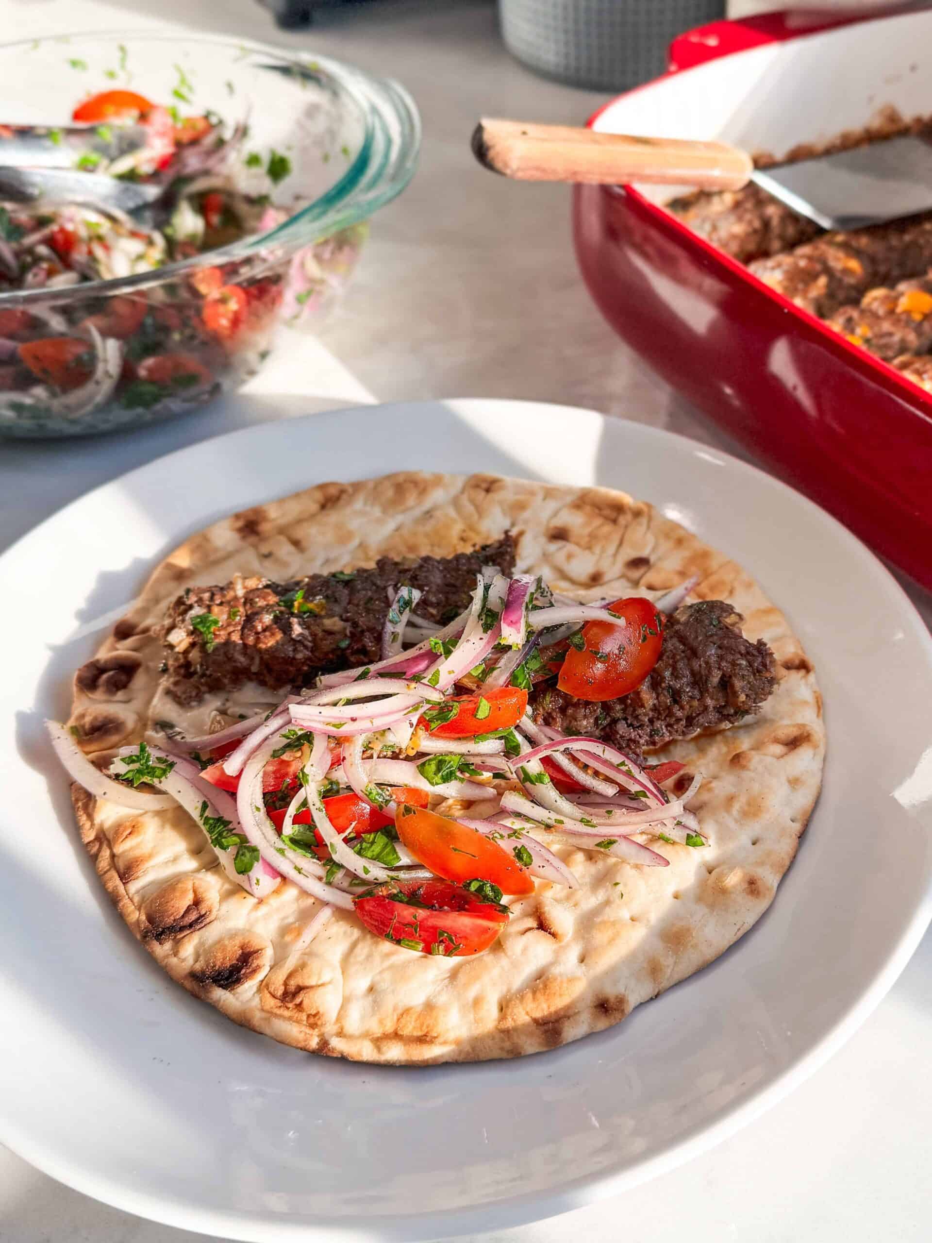 Open ground beef kofta pita sandwich with hummus tomato onion salad and baked kofta visible in the background