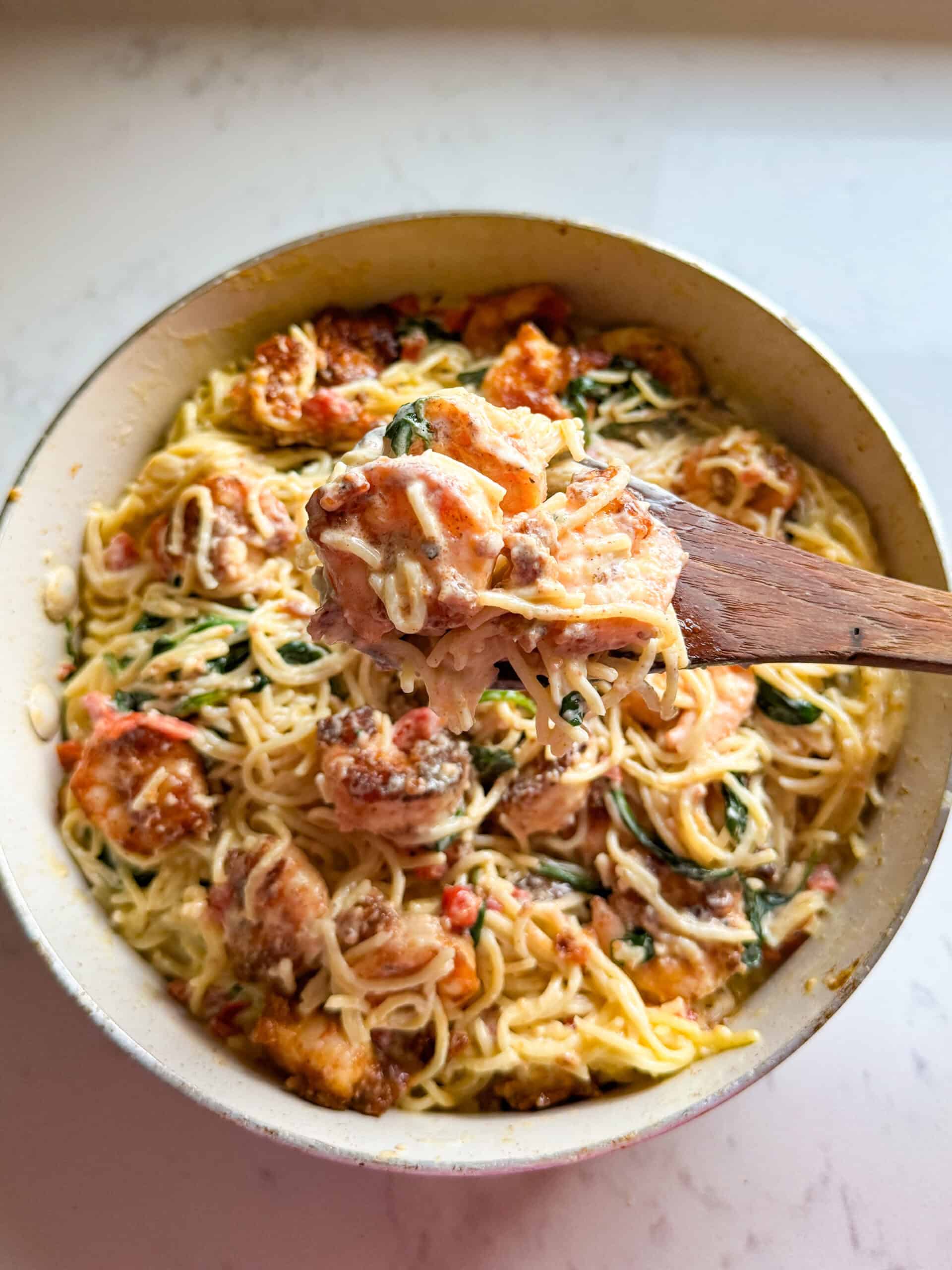 Wooden spoon lifting creamy shrimp pasta with spinach and roasted red peppers
