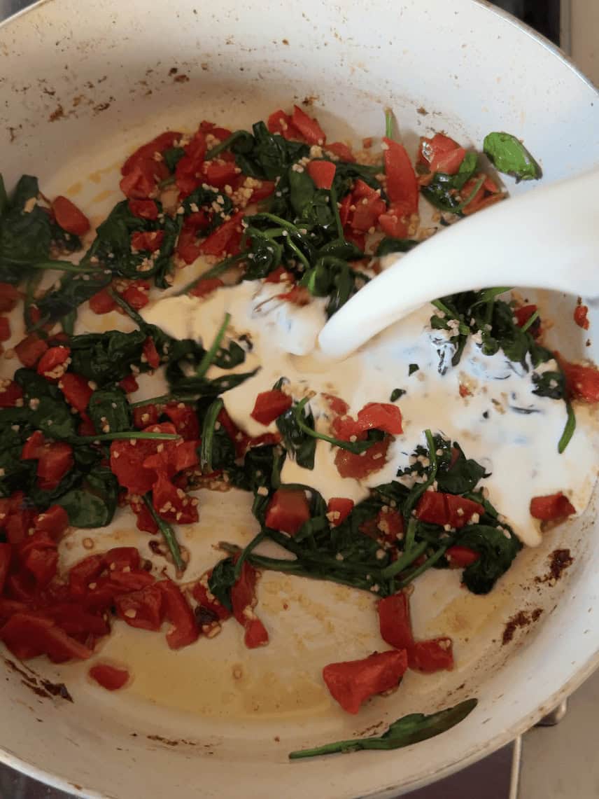 Pouring heavy cream into skillet with roasted red peppers spinach and garlic for shrimp pasta sauce