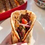 Ground beef kofta pita sandwich with tomato onion salad and hummus on a white plate with oven baked kofta in a casserole dish in the background