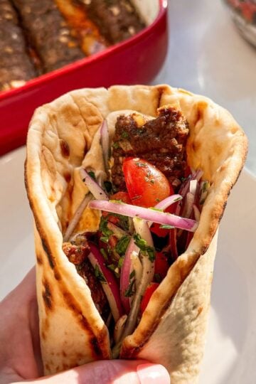 Ground beef kofta pita sandwich with tomato onion salad and hummus on a white plate with oven baked kofta in a casserole dish in the background
