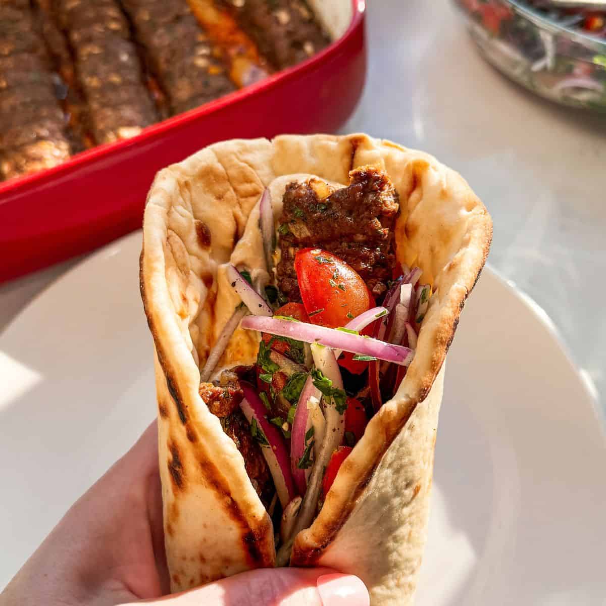 Ground beef kofta pita sandwich with tomato onion salad and hummus on a white plate with oven baked kofta in a casserole dish in the background
