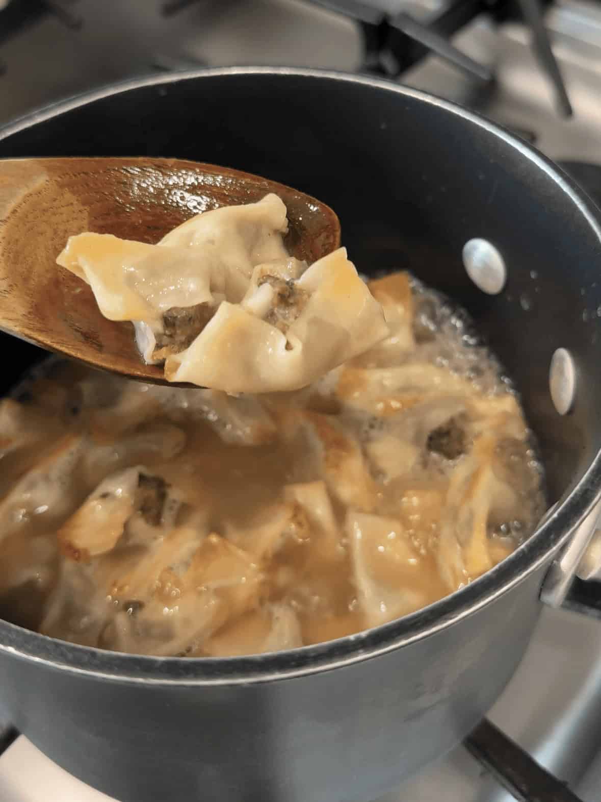 Cooked Armenian Manti dumplings shown in chicken broth with a wooden ladle after simmering.
