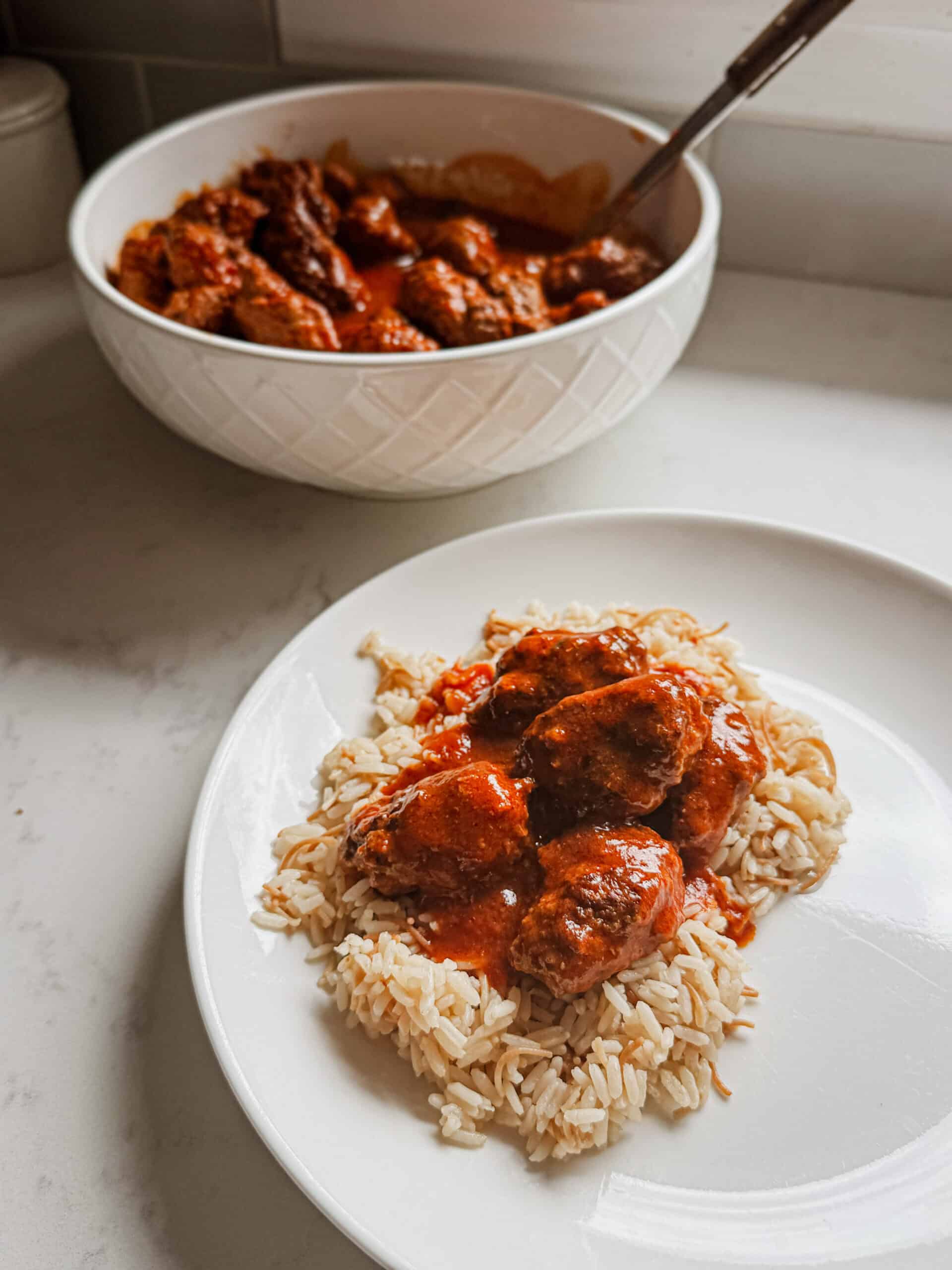 Bowl of sujakaki soutzoukakia Smyrna meatballs in rich tomato sauce served alongside a plated portion over Armenian rice pilaf on white marble
