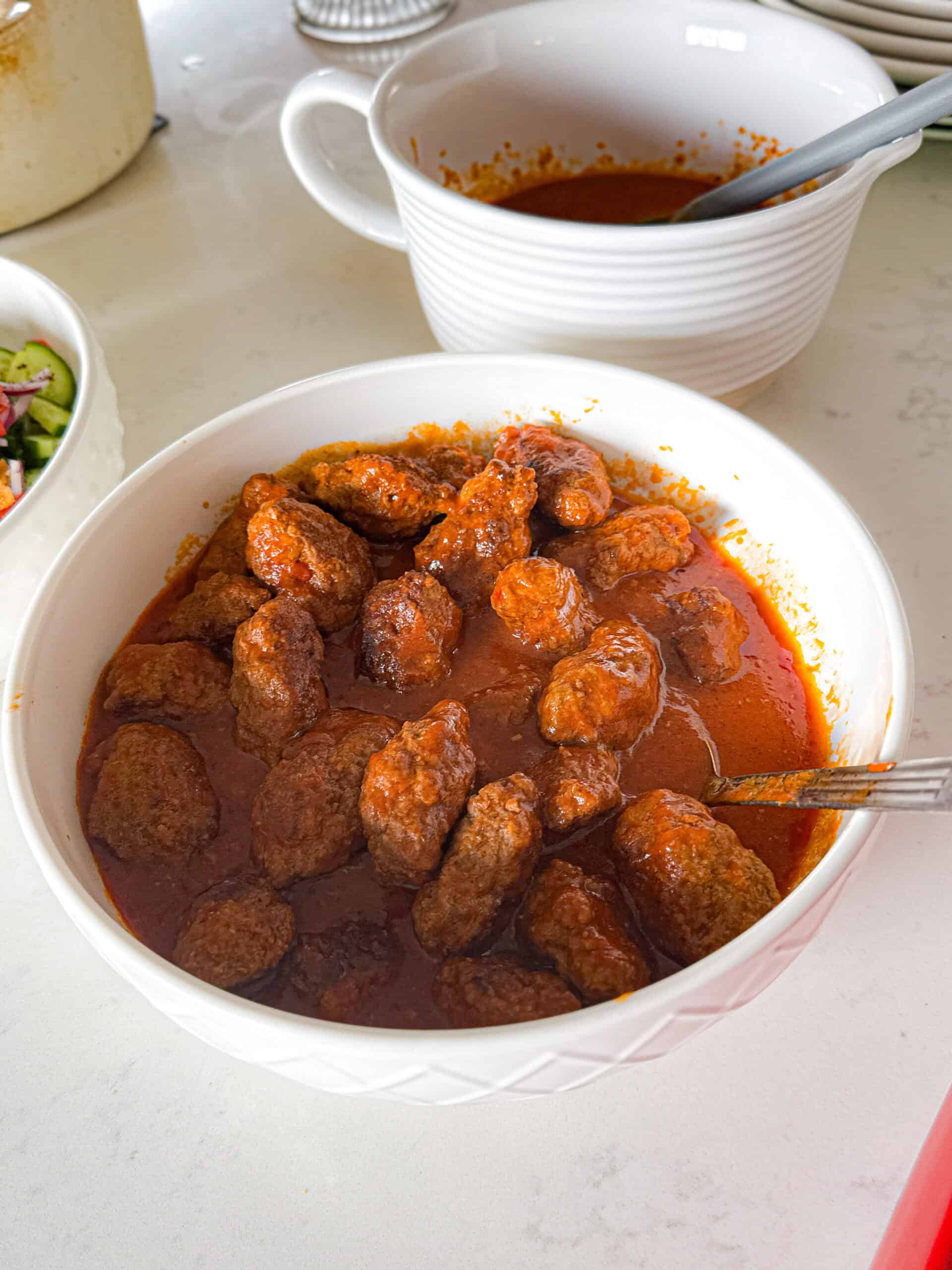 Large white serving bowl filled with sujakaki soutzoukakia Smyrna meatballs in tomato sauce with extra sauce pitcher alongside on white marble