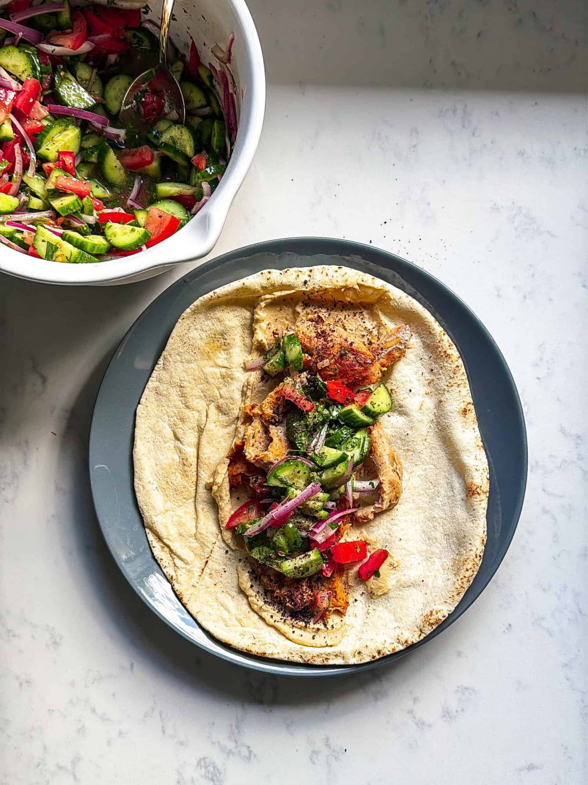 Open chicken doner kabob on flatbread with hummus spread and fresh cucumber tomato red onion salad ready to roll on a gray plate with marble background