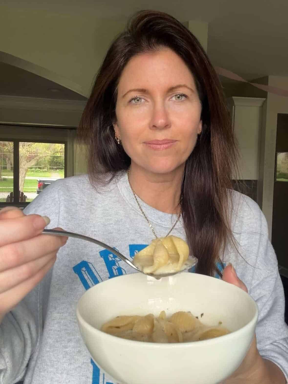 A hand holding a spoon of creamy Armenian yogurt soup with shell pasta over a bowl of Tutmaj Abour