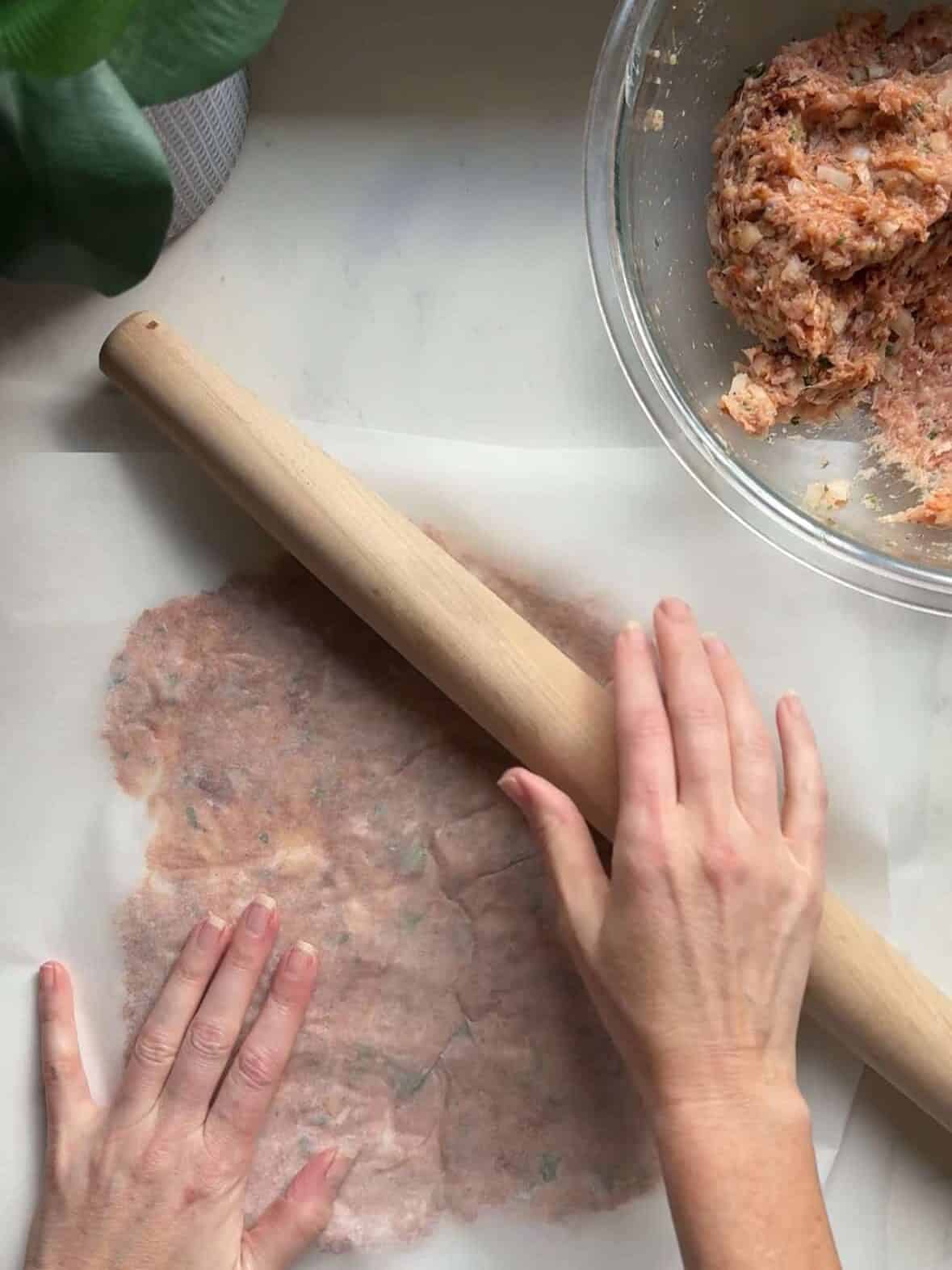 Hands using a rolling pin to flatten seasoned ground chicken doner kabob mixture between parchment paper sheets on a white marble surface