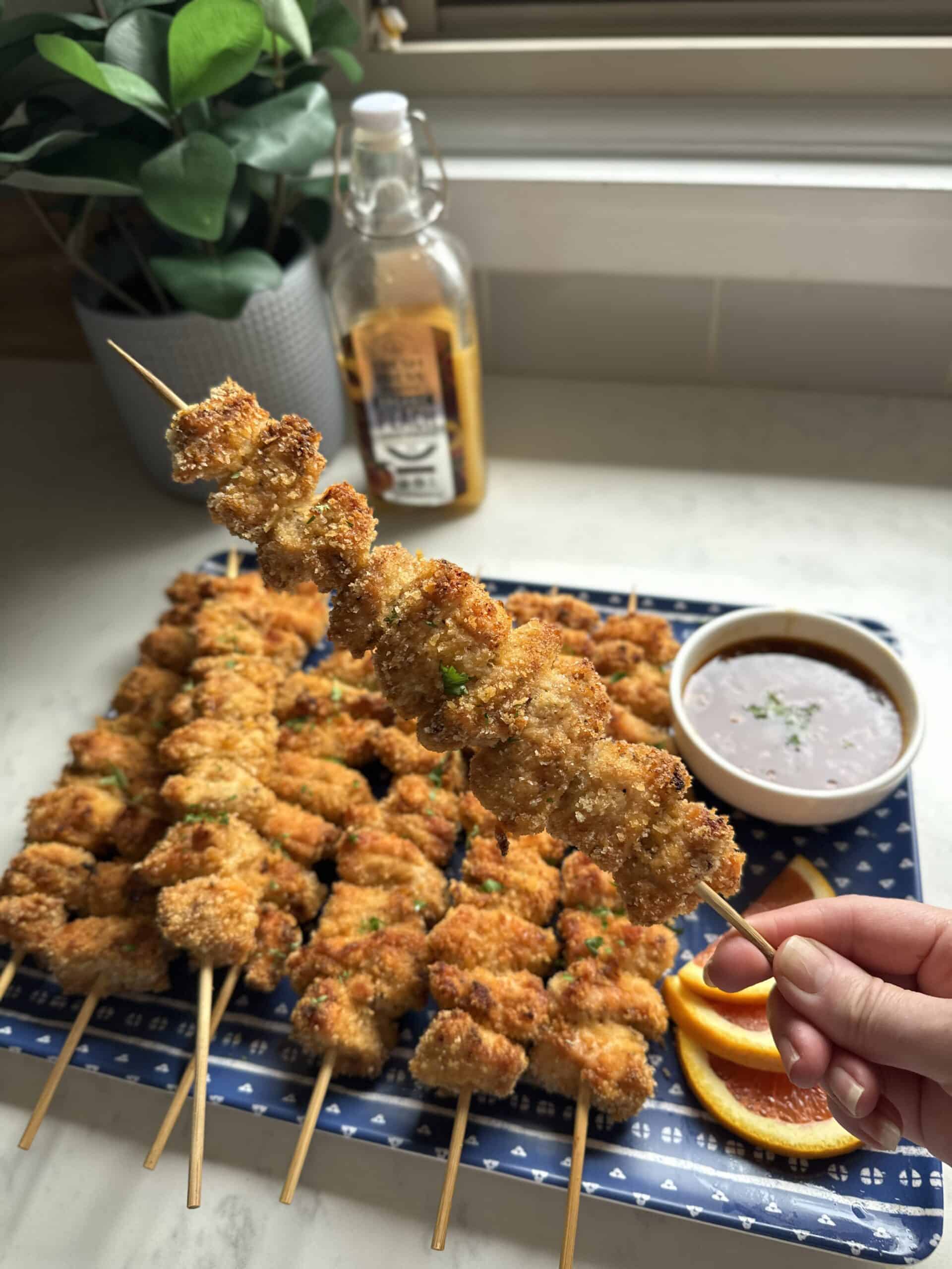 Close-up of a hand holding a single crispy Panko breaded baked chicken skewer over a full platter on a blue tray with Fresh Press Farms Peach Cider Vinegar visible in the background