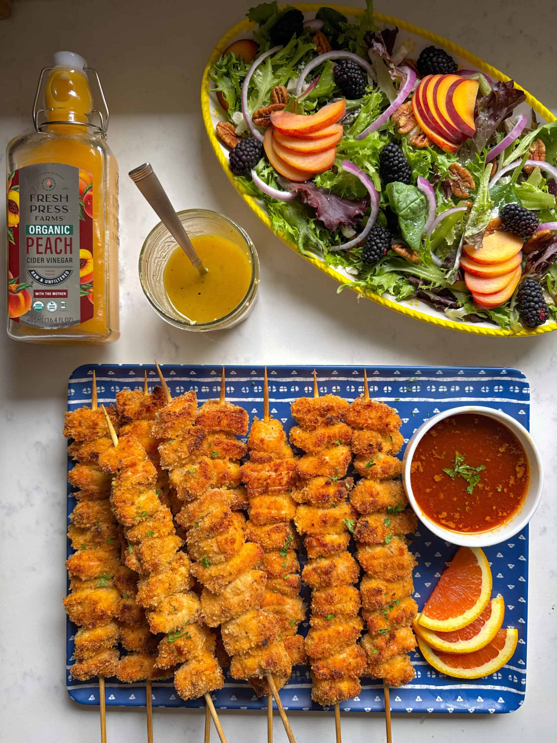 Overhead flat lay of crispy baked chicken skewers on a blue tray alongside a peach summer salad with blackberries, red onion, and pecans in a yellow bowl, with Fresh Press Farms Organic Peach Cider Vinegar bottle and homemade peach dressing