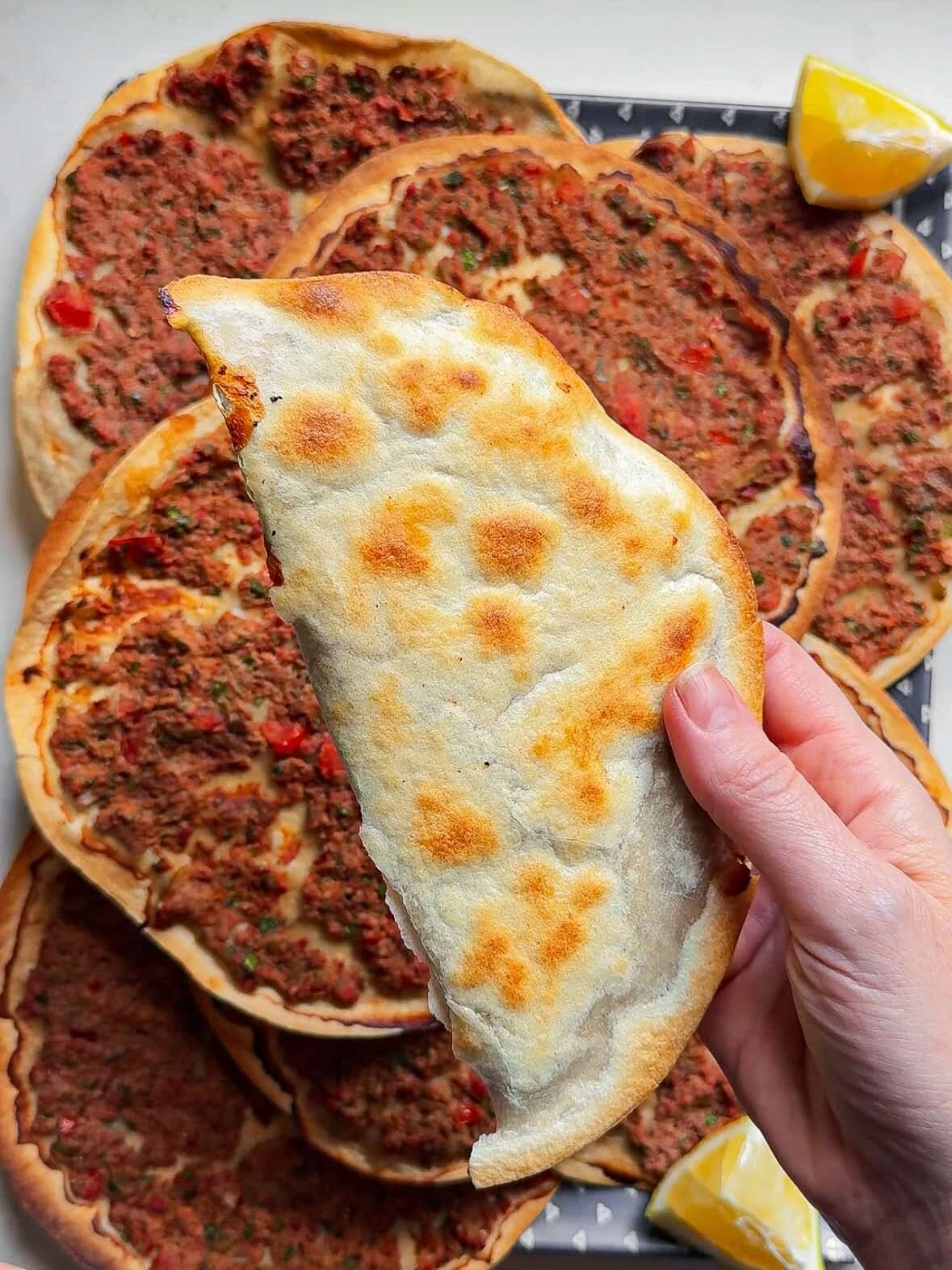 One Armenian lahmajoun folded in half over a stack of lahmajoun on a blue tray with fresh lemon slices, showing the traditional lahmacun wrap serving style.