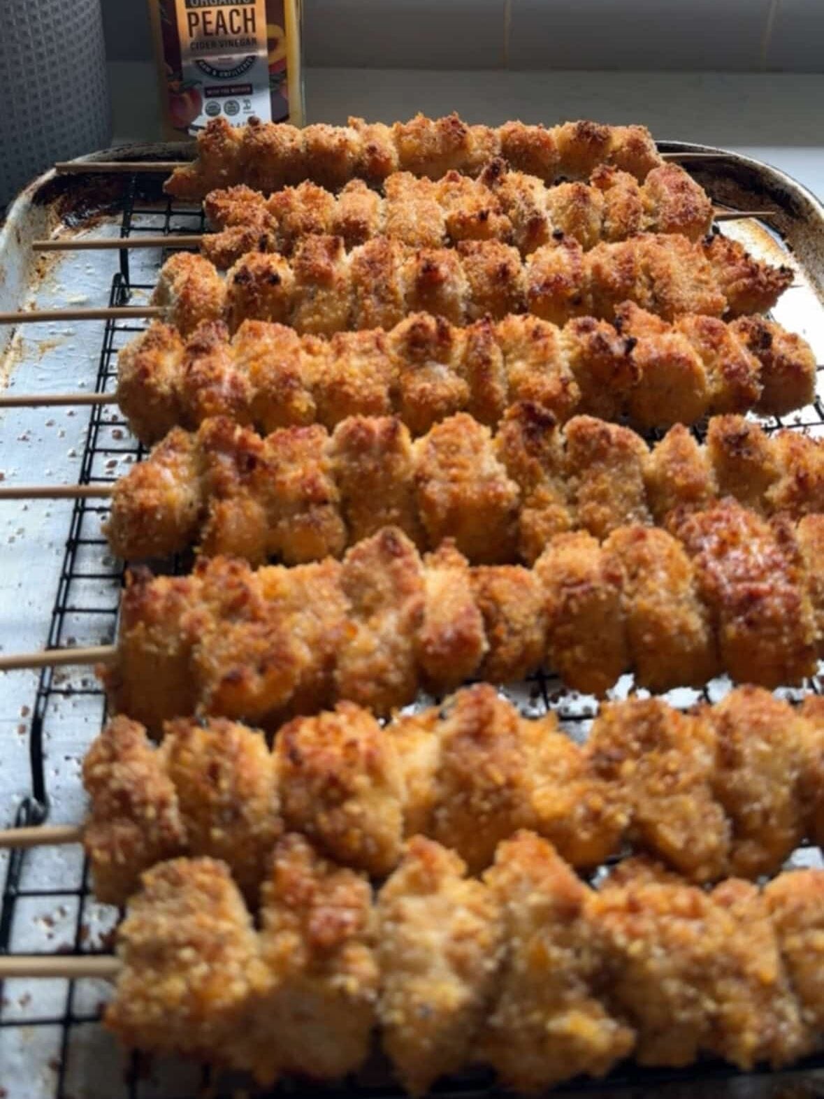 Angled view of golden crispy Panko breaded chicken skewers fresh out of the oven on a wire rack over a baking sheet, with Fresh Press Farms Organic Peach Cider Vinegar bottle in the background