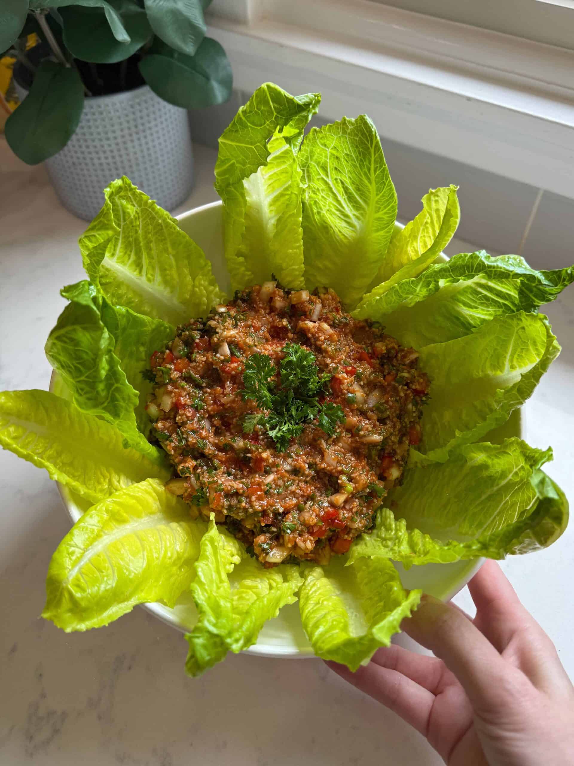 Hand holding a white bowl of Armenian eech red tabouli surrounded by romaine lettuce leaves viewed from above
