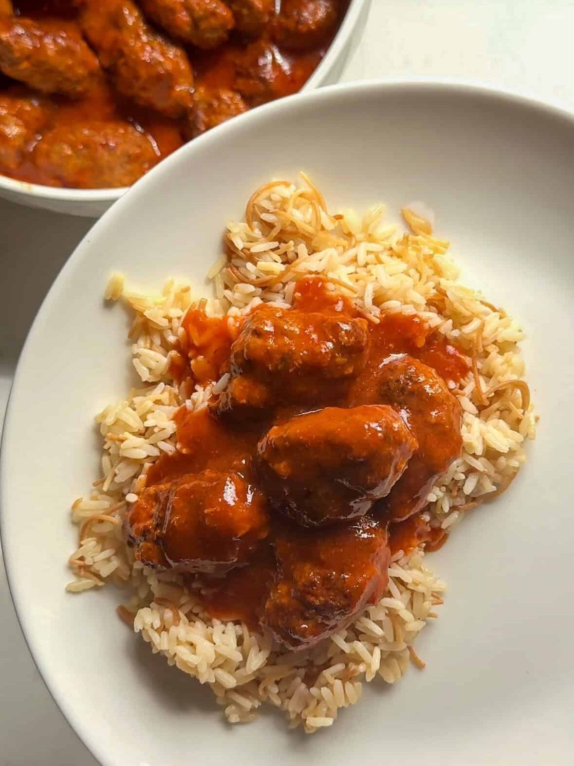 Close up of plated sujakaki soutzoukakia Smyrna meatballs in rich tomato sauce over Armenian rice pilaf on white plate with serving bowl in background