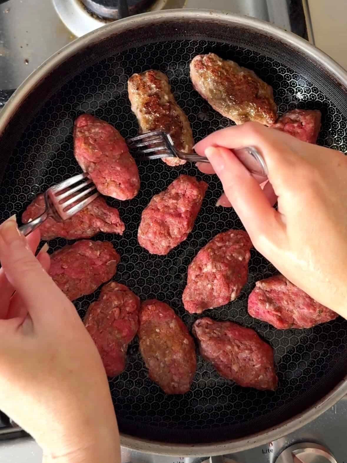 Oblong sujakaki soutzoukakia Smyrna meatballs being seared in a large skillet on gas stovetop with two forks turning them