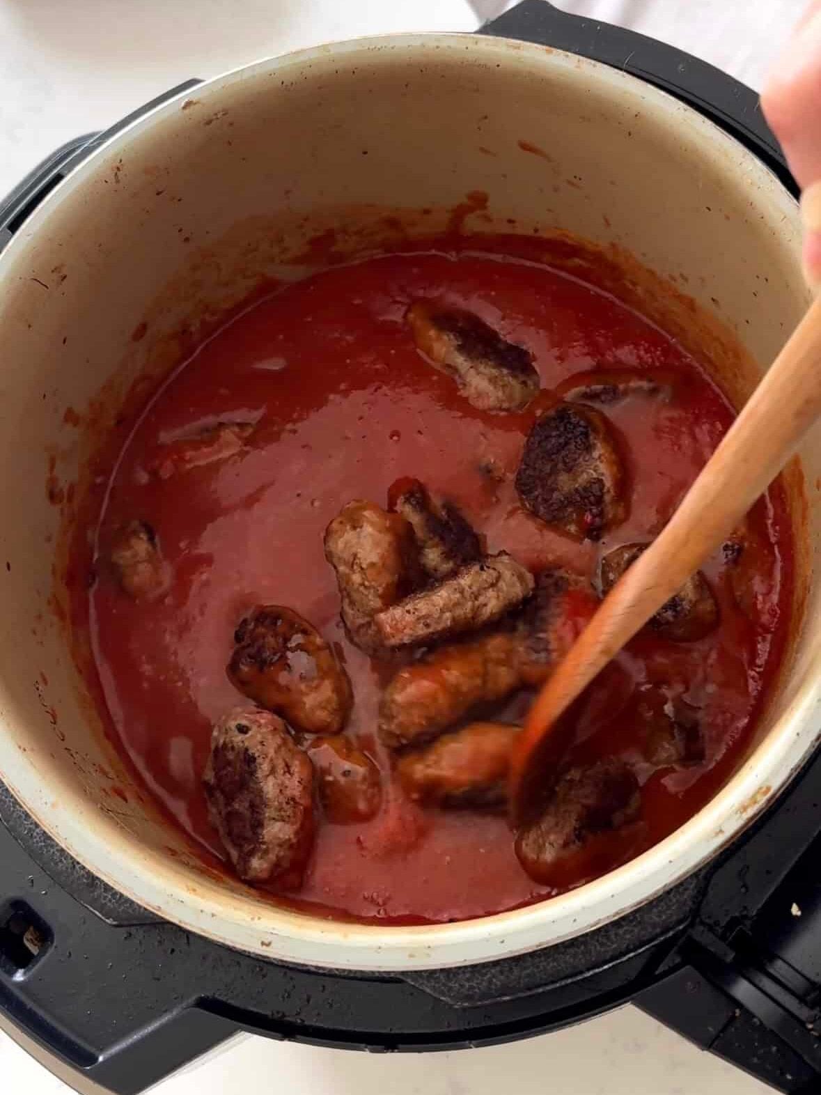 Seared sujakaki soutzoukakia Smyrna meatballs added to Instant Pot pressure cooker with rich tomato sauce being stirred with wooden spoon