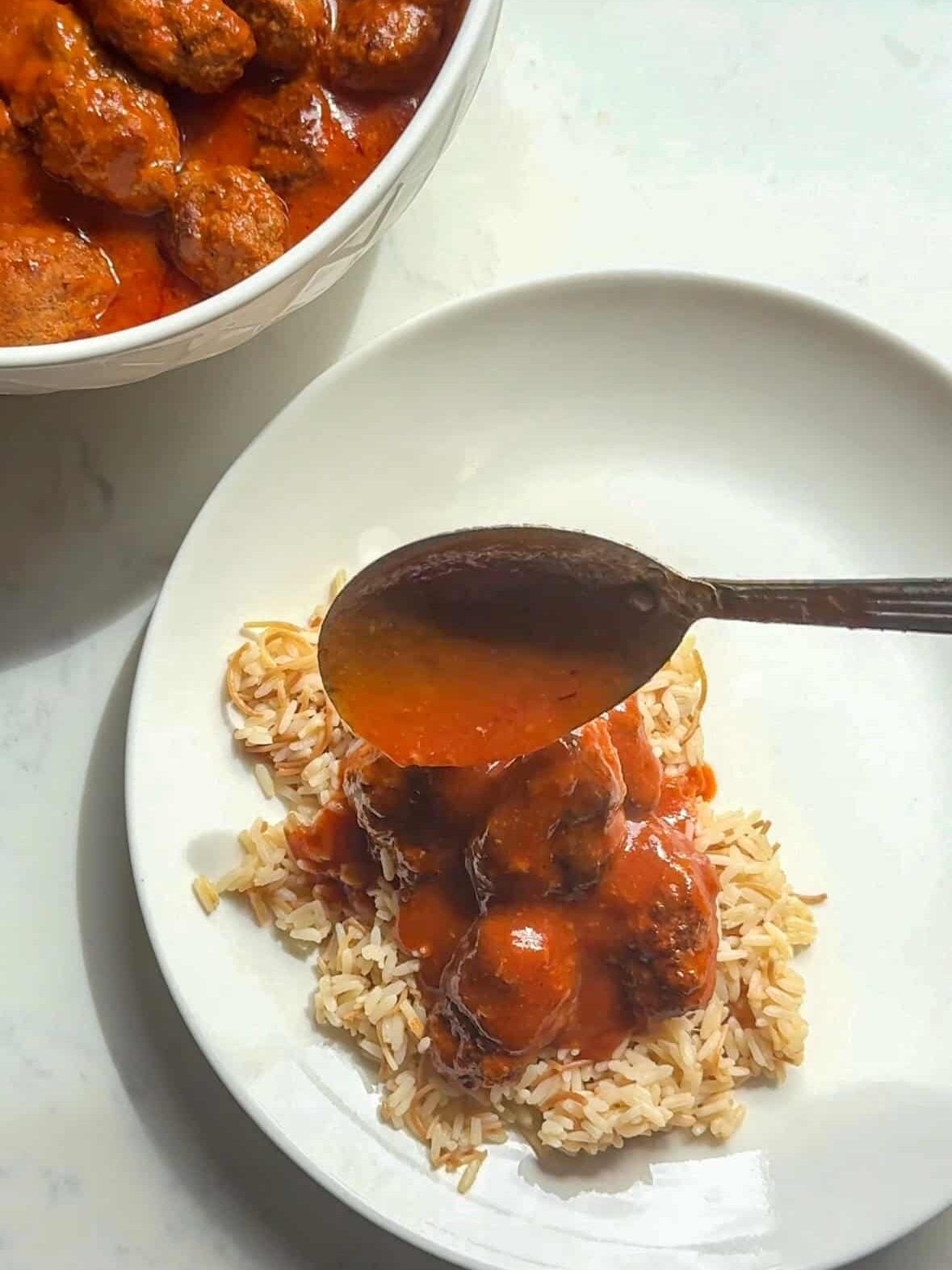 Sujakaki soutzoukakia tomato sauce being spooned over Armenian rice pilaf on white plate with full serving bowl of Smyrna meatballs in background