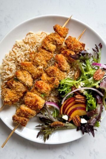 Overhead view of two crispy Panko baked chicken skewers plated on a white plate with toasted rice and a peach summer salad with blackberries, pecans, red onion, feta, and sliced fresh peaches