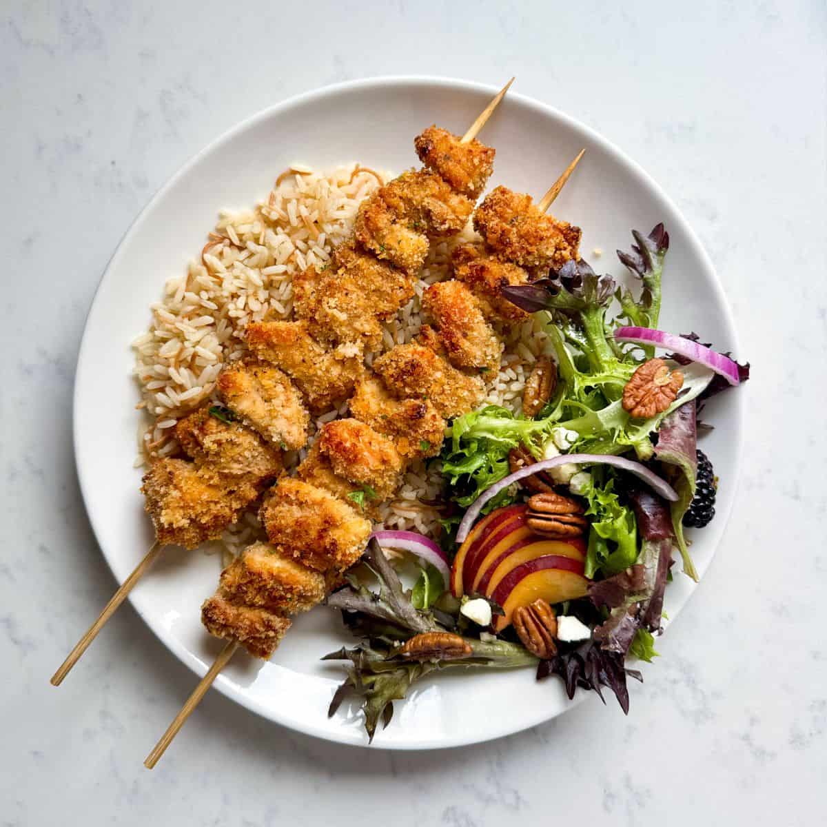 Overhead view of two crispy Panko baked chicken skewers plated on a white plate with toasted rice and a peach summer salad with blackberries, pecans, red onion, feta, and sliced fresh peaches