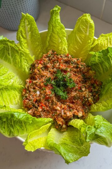 Overhead view of Armenian eech red tabouli in a white bowl surrounded by romaine lettuce leaves on a marble countertop