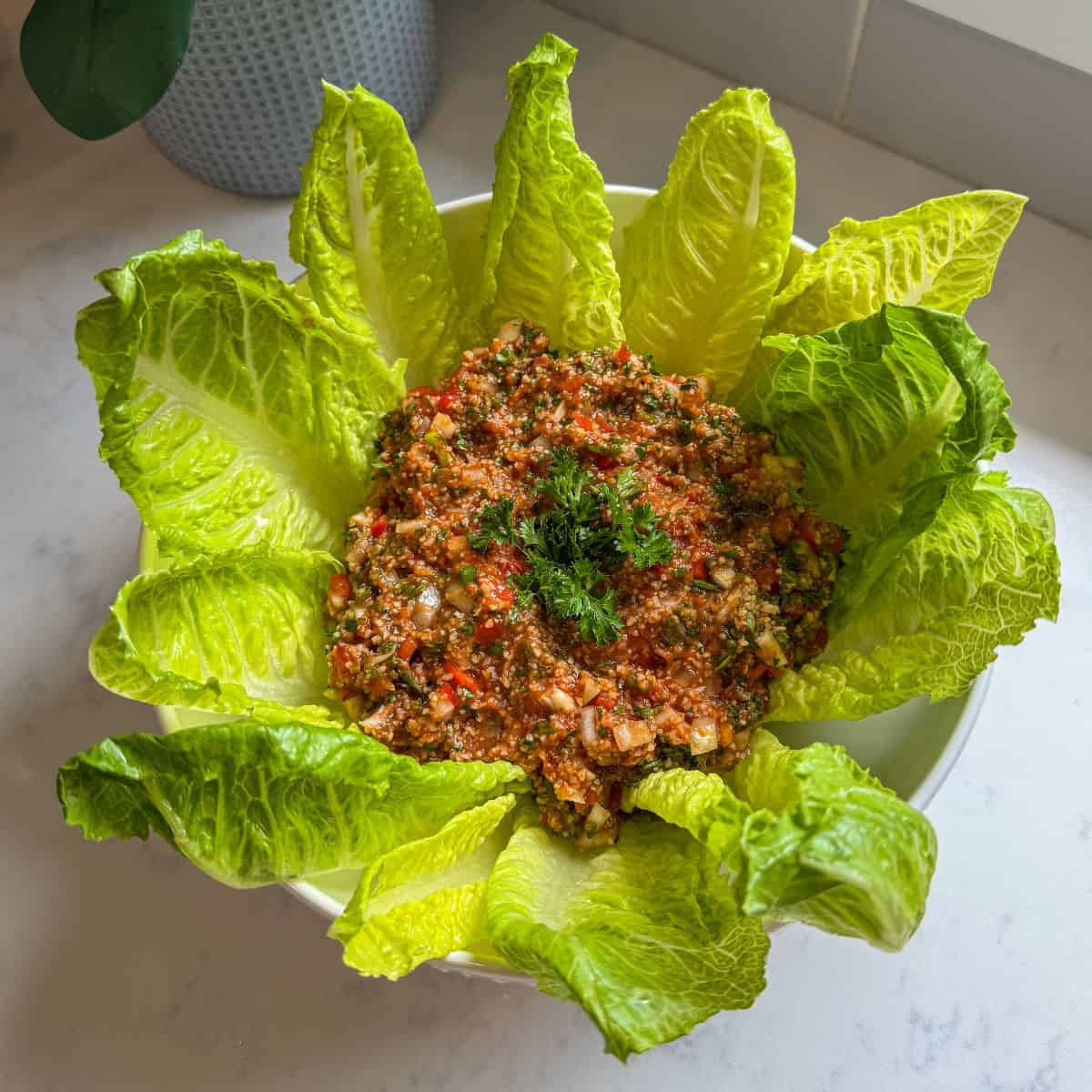 Overhead view of Armenian eech red tabouli in a white bowl surrounded by romaine lettuce leaves on a marble countertop