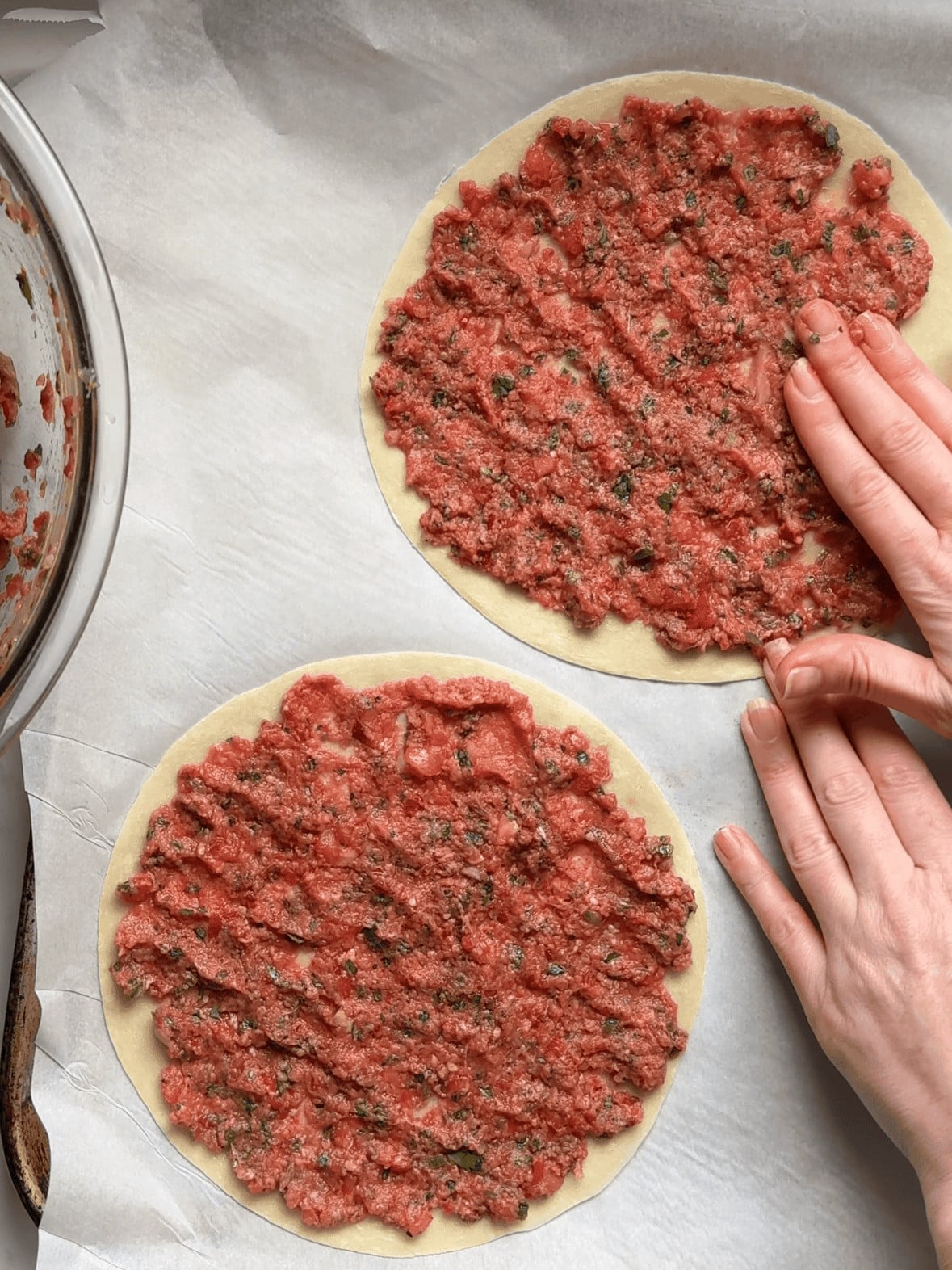 Hands spreading a thin layer of spiced meat mixture evenly across an uncooked flour tortilla for Armenian lahmajoun.