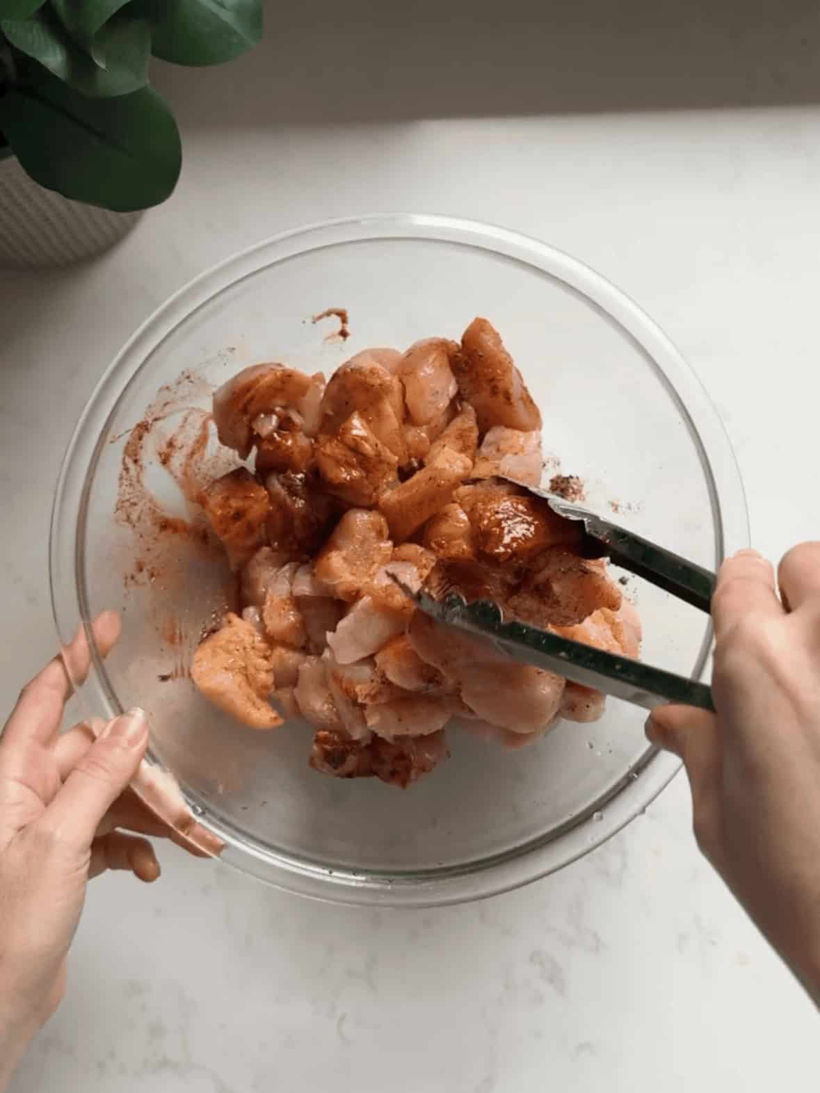 Overhead view of hands using tongs to toss cubed chicken breast in a glass bowl coated with peach cider vinegar marinade with paprika, garlic, and Italian seasoning
