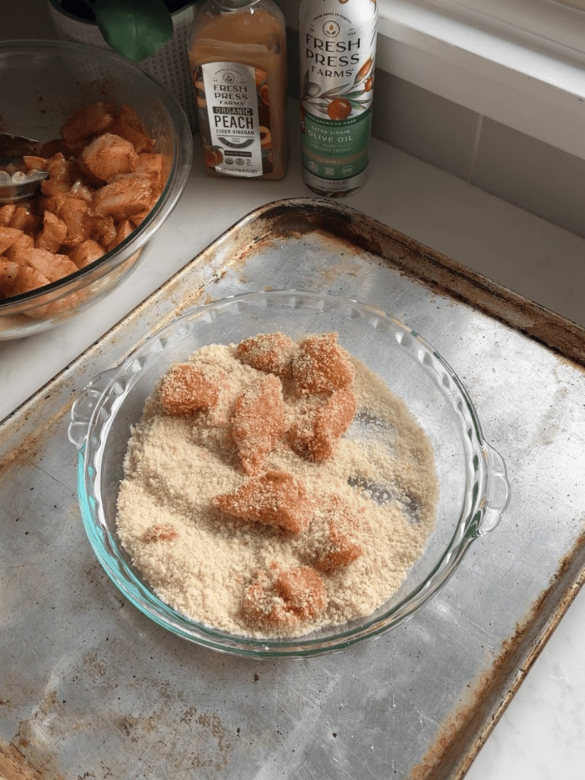 Overhead view of marinated chicken cubes being pressed into Panko breadcrumbs in a glass pie dish on a baking sheet, with Fresh Press Farms Organic Peach Cider Vinegar and Fresh Press Farms olive oil bottles visible in the background